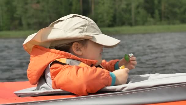 Маленька дівчинка в кепці їсть желе-цукерки, вітрильний каяк на озері — стокове відео