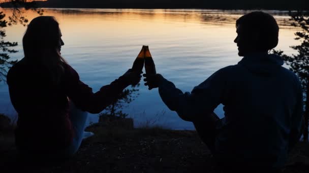 Пара щелкает бутылки пива сидя на берегу спокойной реки — стоковое видео