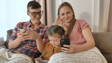 Küçük kız ve ebeveynler kanepeye çağdaş telefonlar koyarlar.