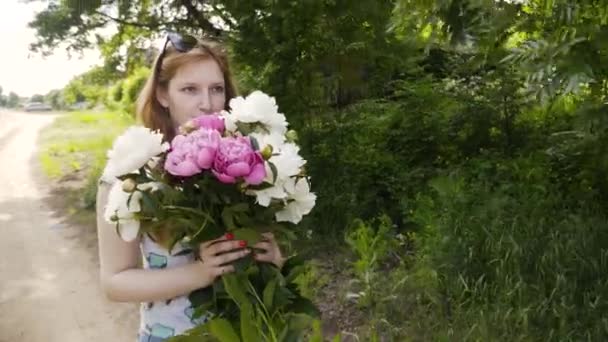 ピオニの花束を持つ魅力的な女の子は 村の道路上にあります 赤と白のピオネの花束を持つ女の子 — ストック動画