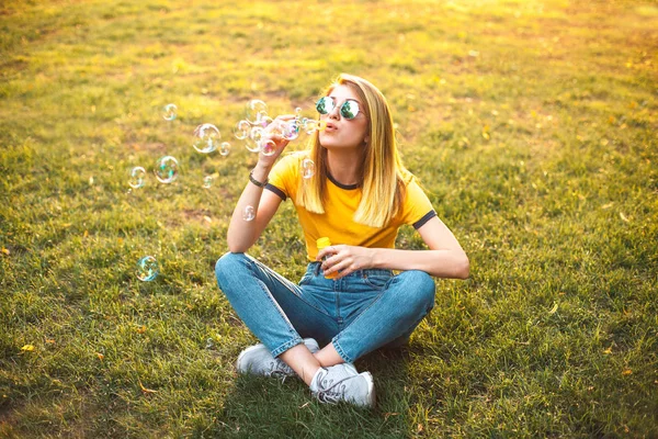 çimenlerin üzerinde oturan ve parkta sabun köpüğü üfleme genç şık kadın