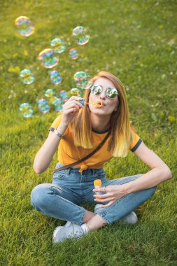 Genç bir kadın çim üzerinde oturur ve sabun kabarcıkları darbeler.