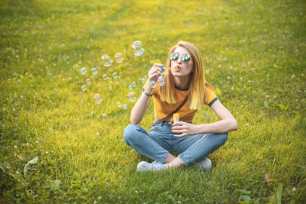 一个戴着太阳镜的年轻女子坐在草坪上吹泡肥皂泡 — 图库照片