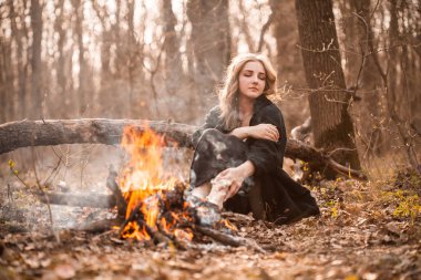Attractive young woman with black cape sitting in forest near bonfire. clipart