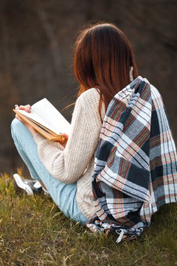 Genç bir kadın dışarıda oturup kitap okuyor. Geri görünüm