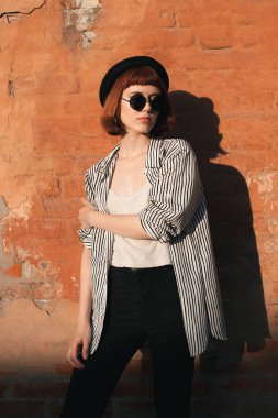 Çizgili gömlekli, şapkalı ve güneş gözlüklü genç manken turuncu duvarın yanında poz veriyor.