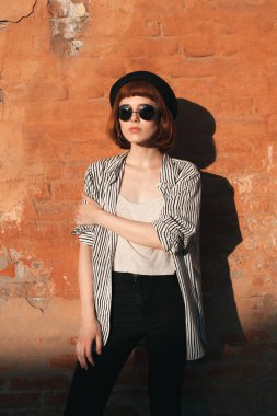 Çizgili gömlekli, şapkalı ve güneş gözlüklü genç manken turuncu duvarın yanında poz veriyor.