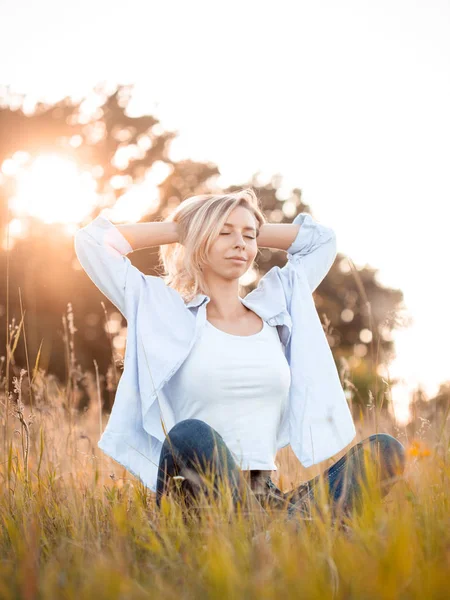 Genç sarışın kadın gün batımında çimlerin üzerinde oturuyor. Gözler kapalı.