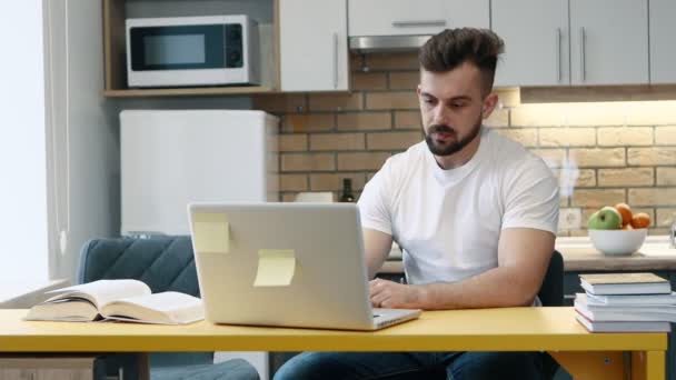 Homem aprende e escreve trabalho em um computador em projeto usando um livro didático sentado na cozinha em casa . — Vídeo de Stock
