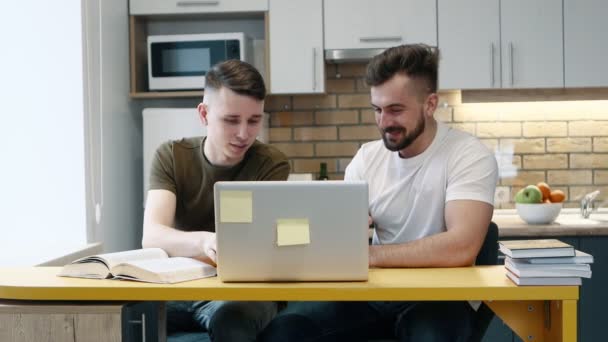 Two guys learn and work on a computer on project using a textbook sitting in the kitchen at home. Talking each other. — Stock Video