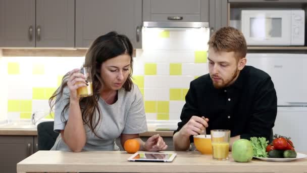 年轻夫妇的男人和女人与平板电脑吃早餐坐在厨房里的家. — 图库视频影像