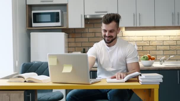 El hombre aprende y escribe el trabajo en una computadora en proyecto usando un libro de texto sentado en la cocina en casa . — Vídeo de stock