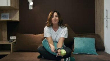 TV izlerken, yatak odasında yatağın üstüne oturan bir elma yemek kadın. Anahtarları kanalları uzaktan kumanda ile.