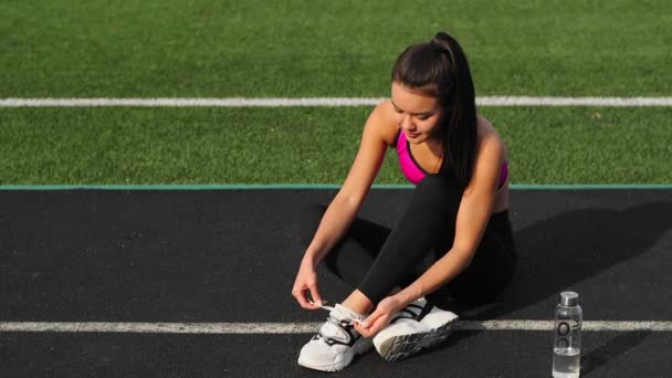 Deportiva asiática chica en ropa deportiva es atando cordones en zapatillas de deporte sentado en el estadio antes de entrenar . — Vídeo de stock