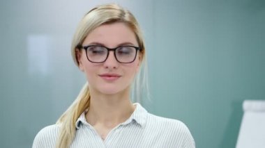 Ofis kıyafetleri ve gözlüklü genç sarışın iş kadını kameraya bakıyor ve gülümsüyor..