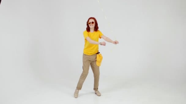 Grappig roodharige jong meisje met gele ballon in de hand is dansen met een populaire bewegingen op lichte achtergrond. — Stockvideo