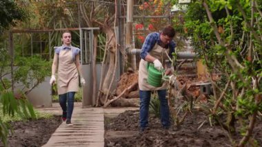 Bahçıvanlar serada çalışıyor, adam bitkileri sulıyor, kadın yolda yürüyor.
