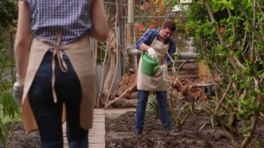 Bahçıvanlar serada çalışıyor, adam bitkileri sulıyor, kadın yolda yürüyor.