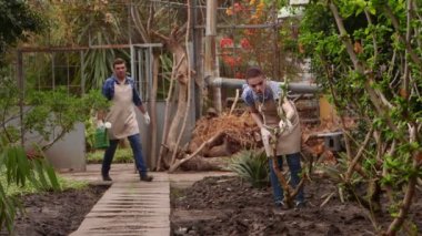 Bahçıvanlar serada çalışıyor, adam bitkileri sulıyor, kadın yolda yürüyor.