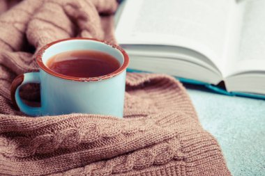 Bir fincan sıcak çay, açık kitap ve sıcak örülmüş battaniye.