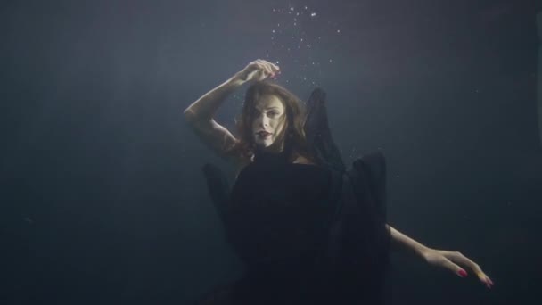 Young woman in black dress swimming underwater on dark background — Stock Video