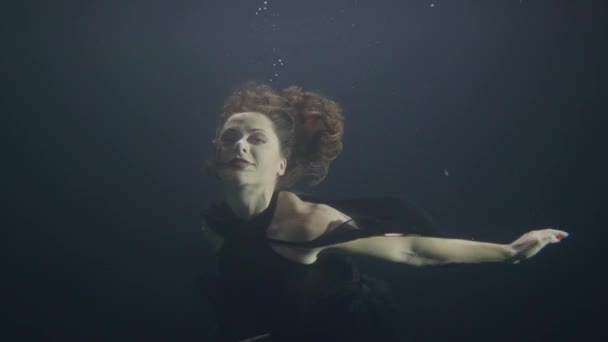 Hermosa mujer en vestido negro nadando bajo el agua en la piscina sobre fondo oscuro — Vídeos de Stock