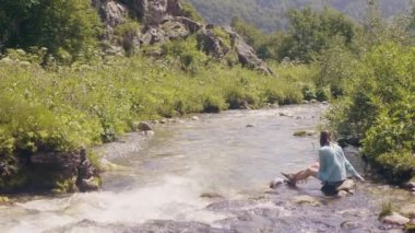 Taşlı nehir kıyısında oturan ve doğa yürüyüşü sırasında dinlenme mutlu kadın.
