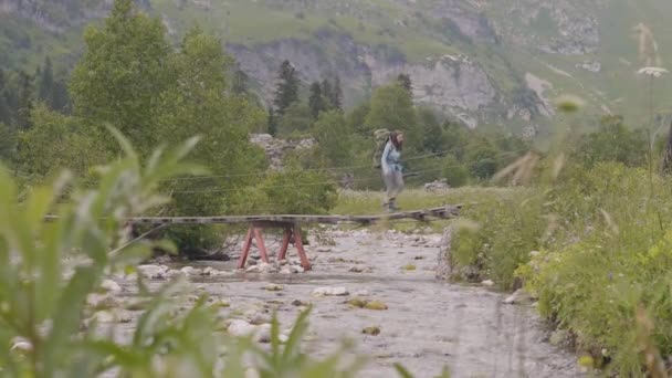 Turistas com mochilas andando na ponte suspensa sobre o rio da montanha — Vídeo de Stock