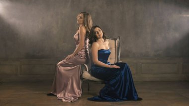 İki kız şarkıcı bir sandalyede oturmak ve şarkı. 4 k longshot. Lüks mavi ve pembe akşam elbiseler. Sarışın ve esmer kadın.