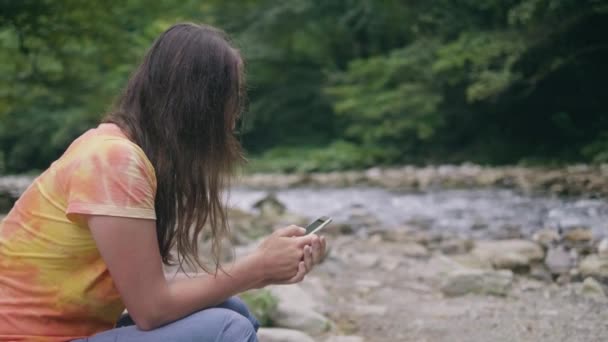 Mujer joven navegando por el teléfono móvil mientras descansa en la orilla del río — Vídeos de Stock