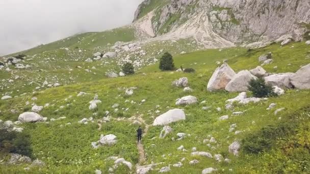 Женщина с видом на дрон ходит по горной тропе среди больших камней по зеленой траве — стоковое видео