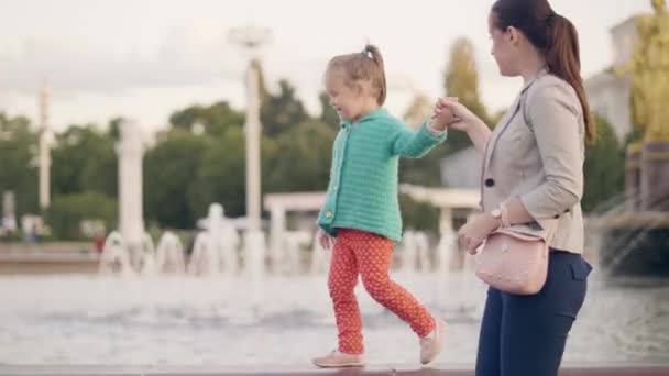 夏の公園の噴水のエッジの上を歩く幼い娘と幸せママ ママと娘の夏の日を一緒に過ごすと 噴水の背景に都市公園を歩いて — ストック動画