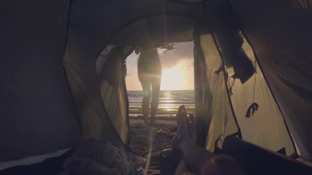 Vista da silhueta de tenda turística jovem na paisagem dourada do pôr do sol no mar e pés masculinos deitados dentro. Ponto de vista homem descansando na tenda do acampamento e mulher silhueta no amanhecer fundo . — Vídeo de Stock