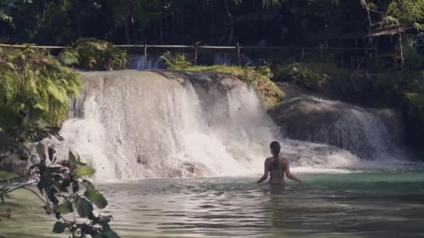 Mujer joven bañándose en cascada en el bosque tropical y chorro de agua salpicando en el fondo. Agua de flujo que vierte sobre piedras en el río de montaña desde la cascada en el bosque selvático . — Vídeo de stock