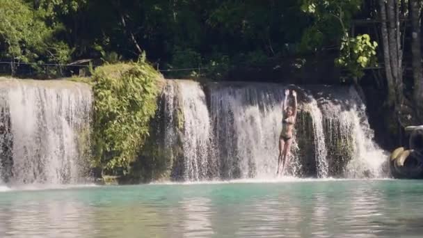Junge Frau springt in Bergsee vom Seil und plätschert Wasserfall auf Hintergrund. Zeitlupe. glückliche Frau, die in Wasserfallsee von Seil und Stock springt. Extreme Ruhe im Sommerurlaub. — Stockvideo