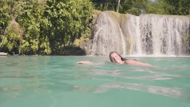 Mujer sonriente nadando en el lago de la cascada en la selva tropical. Mujer alegre disfrutando de nadar en aguas cristalinas desde la cascada de montaña. Chica en el lago de montaña y salpicaduras de agua de fondo . — Vídeos de Stock