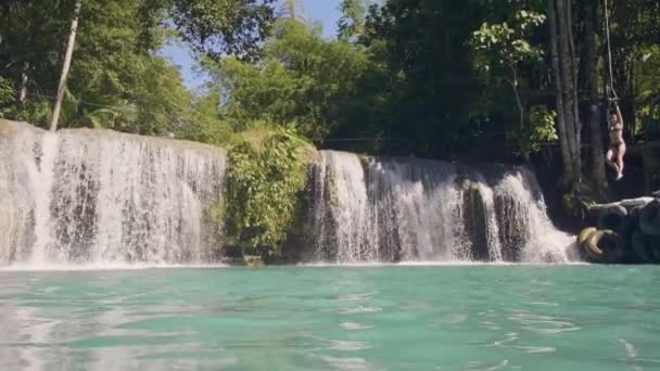 Mladá žena skákat v jezeře vodopád z lana a stick zpomaleně. Veselá žena skákat z lana v modré vodě z tekoucí vodopád v deštném pralese. — Stock video