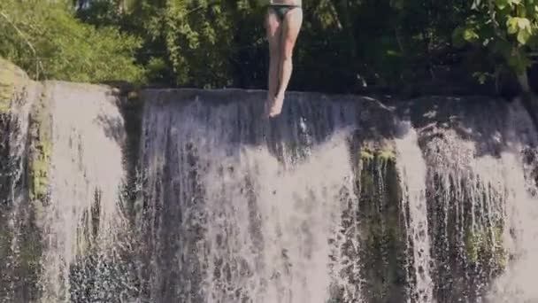 Junge Frau springt in Bergsee vom Seil und plätschert Wasserfall auf Hintergrund. Zeitlupe. glückliche Frau, die in Wasserfallsee von Seil und Stock springt. Extreme Ruhe im Sommerurlaub. — Stockvideo