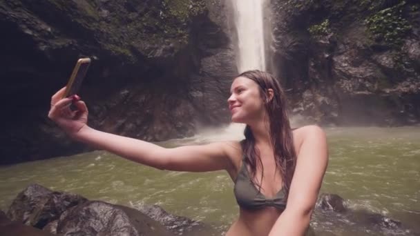 Young woman travel blogger doing selfie photo to mobile phone on mountain waterfall background. Smiling woman shooting mobile selfie on splashing waterfall flowing in rainforest. — Stock Video