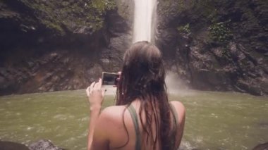 Gülümseyen kadın yağmur ormanlarında cep telefonuna tropik şelale panoramik fotoğraf çekim. Tropikal orman şelale, yolculuk sırasında fotoğraf yapıyor blogger seyahat
