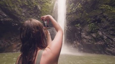 Şelale orman orman içinde sıçramasına fotoğraf için akıllı telefon kullanan gezgin kadın. Yağmur ormanlarında yürüyüş sırasında cep telefonu video tropik şelale çekim Kadın turist.