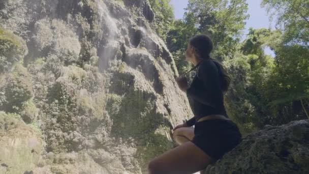 Mujer joven sentada en las rocas mirando la cascada que fluye desde el acantilado en el bosque tropical. Mujer turística disfrutando del paisaje de cascada mientras viaja en la selva tropical. Naturaleza salvaje en la selva . — Vídeos de Stock