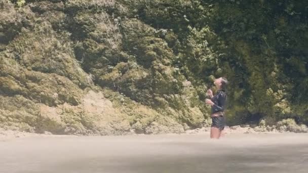 Tourist woman standing in mountain lake under water drops falling from waterfall in rainforest. Happy woman enjoying fresh water flowing from tropical waterfall — Stock Video