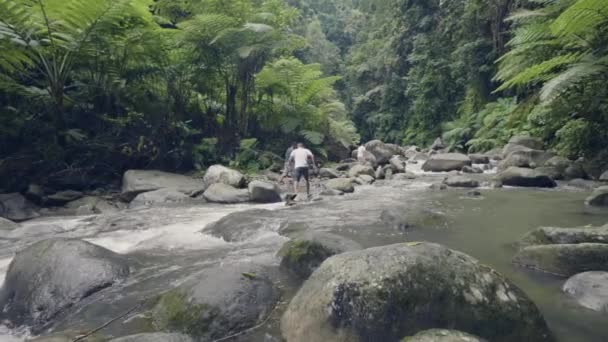 Grupp turist människor gå på klipporna i berg flod som rinner i regnskogen. Drone Visa personer som reser på stony river i djungeln skogen på Bakgrund tropiska palmer och växter. — Stockvideo
