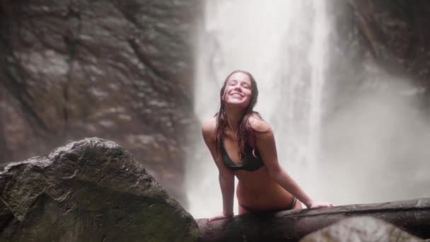 Brunett kvinna med blött hår och bikini leende och poserar främre kameran på flödande mountain vattenfall på bakgrunden. Lycklig kvinna ser att kameran på stänk vattenfall landskap i regnskogen. — Stockvideo
