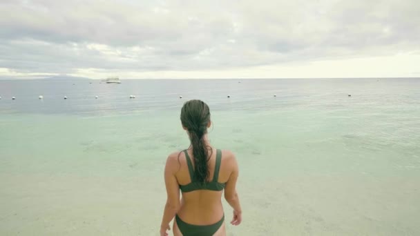 Mujer delgada en traje de baño que entra al agua de mar en la playa de verano en el fondo nublado del cielo. Mujer turística nadando en el océano durante las vacaciones de verano en la playa del complejo . — Vídeo de stock