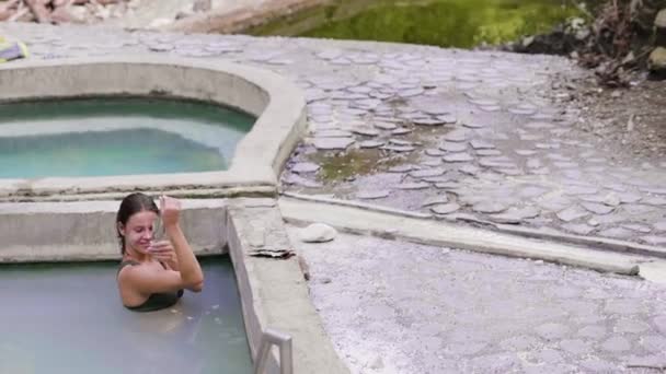 Beautiful woman applying mud mask on face and body skin while taking mineral bath in natural resort. Happy woman enjoying thermal bath in water pool in outdoor spa. — Stock Video