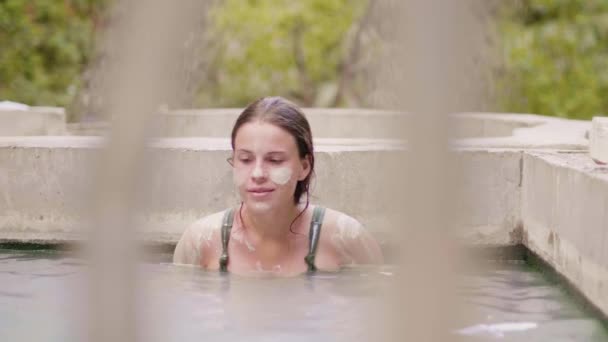 Mujer joven con máscara de barro en la cara de la piel relajante en el barro mineral en el spa al aire libre. Hermosa mujer nadando en baño termal. Terapia de spa natural . — Vídeos de Stock