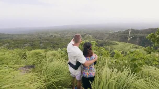 Pareja de turistas mirando la montaña y las tierras altas cubierto de bosque verde en el horizonte de fondo. Pareja viajera abrazándose en una colina verde y disfrutando del paisaje de montaña . — Vídeos de Stock