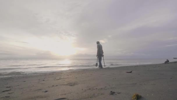 ヘッドフォンの男彼の犬と海砂のビーチで金属探知機の作品では、中年時日の出 — ストック動画